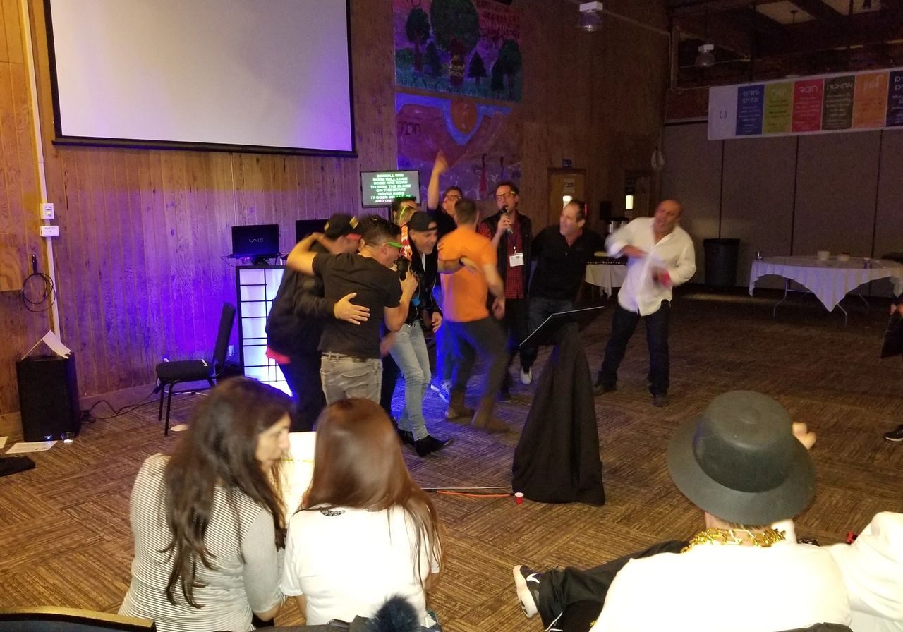 karaoke dj, los angeles dj A group of people gather on a stage under a projector screen, some hugging and celebrating, while others sit and watch in a warmly lit room with wood-paneled walls.