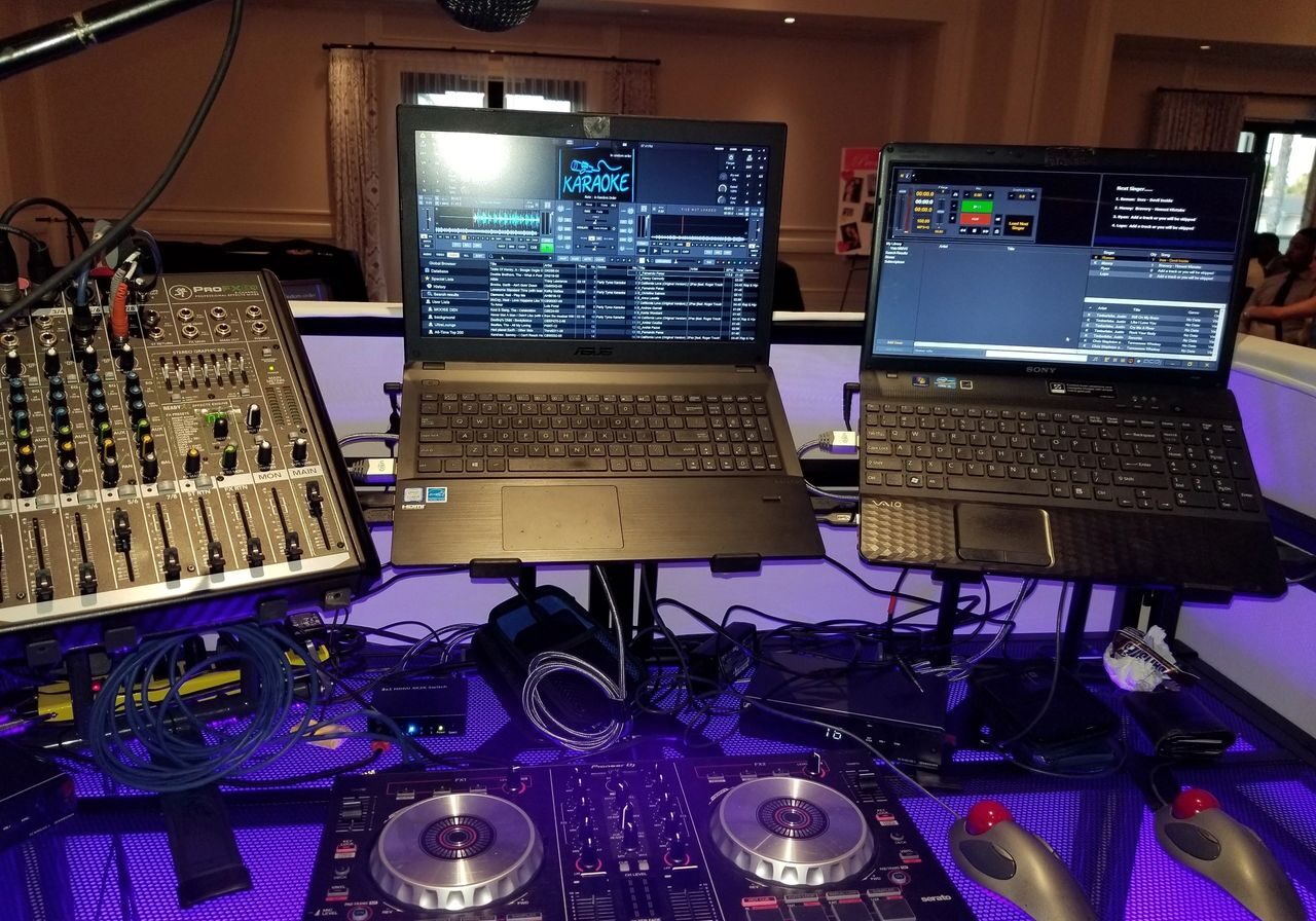 DJ and karaoke setup with a mixer, two laptops displaying DJ and karaoke software, a microphone, cables, and a DJ controller on a lit table.
