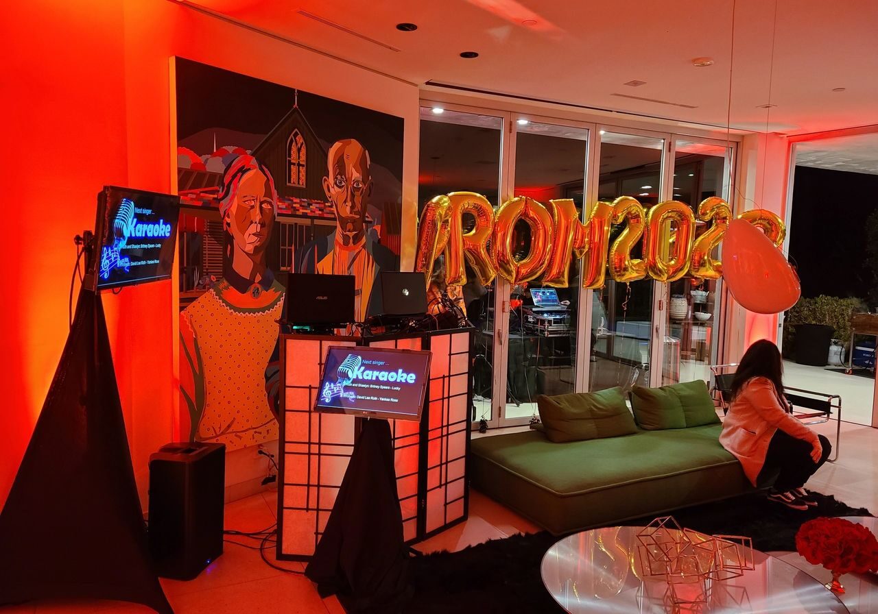 AirBnB rental A living room decorated with "PROM 2023" gold balloons, a karaoke setup, colorful lighting, and a mural; a person sits on a green couch.