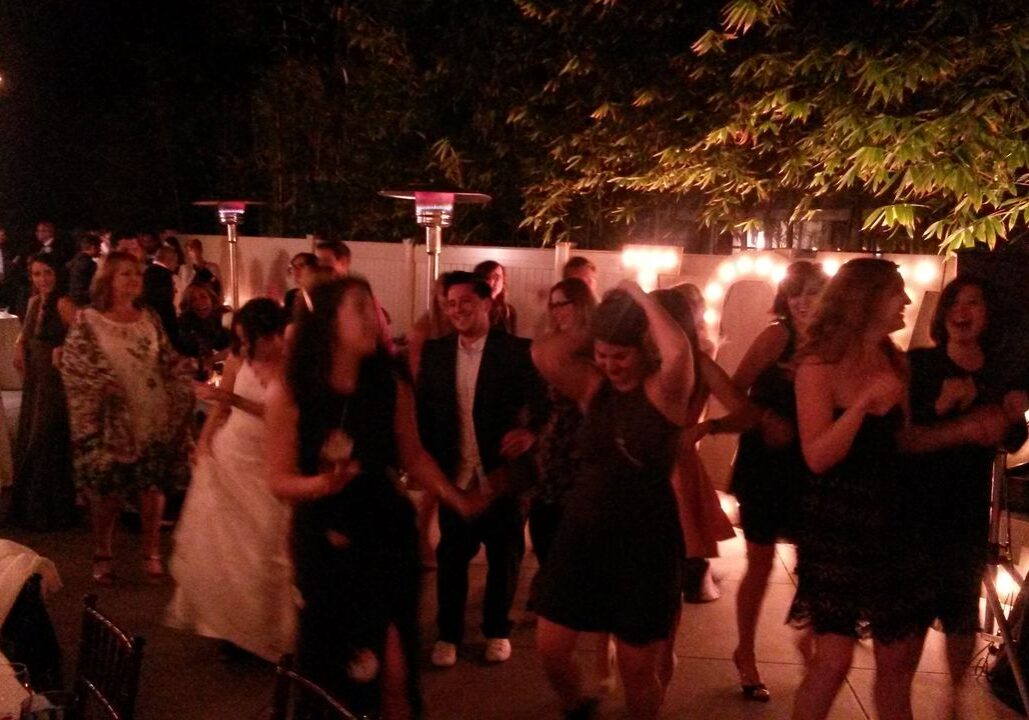 A group of people dance and socialize at an outdoor evening event, with string lights and patio heaters visible in the background.