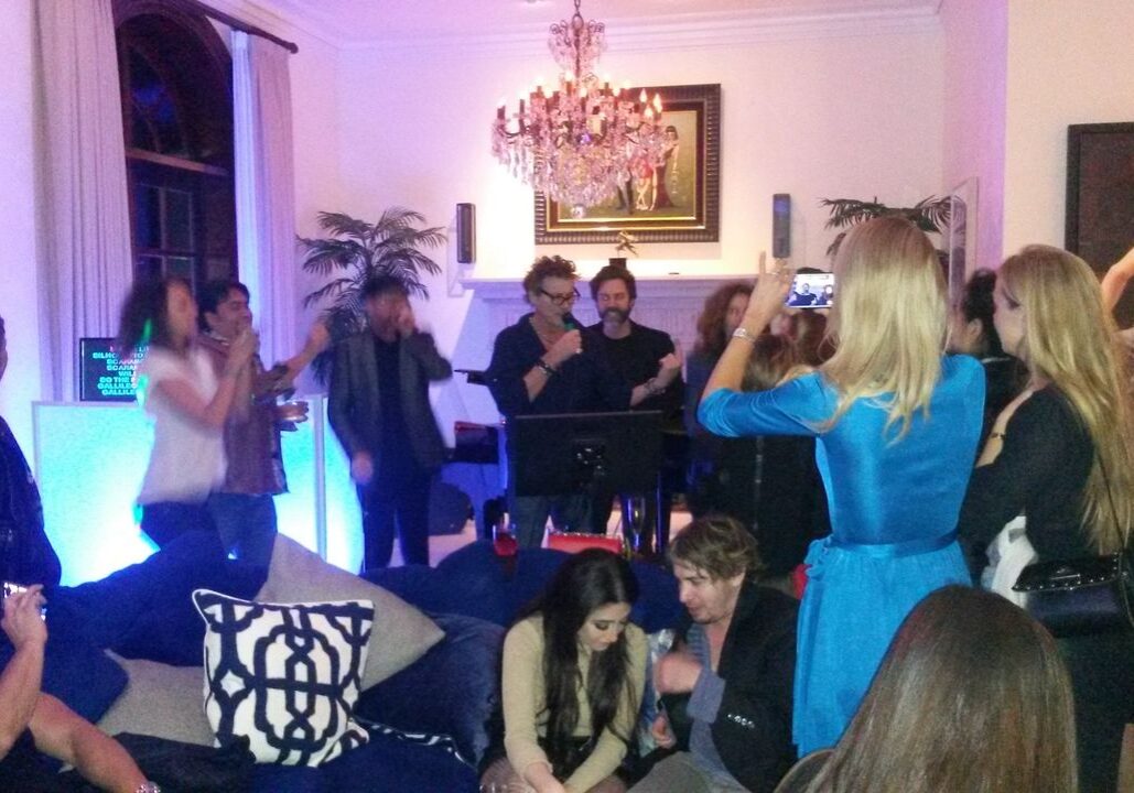 A group of people gathers in a living room, some standing and others sitting, with colorful lighting and a chandelier overhead. A woman in blue is taking a photo of the crowd.