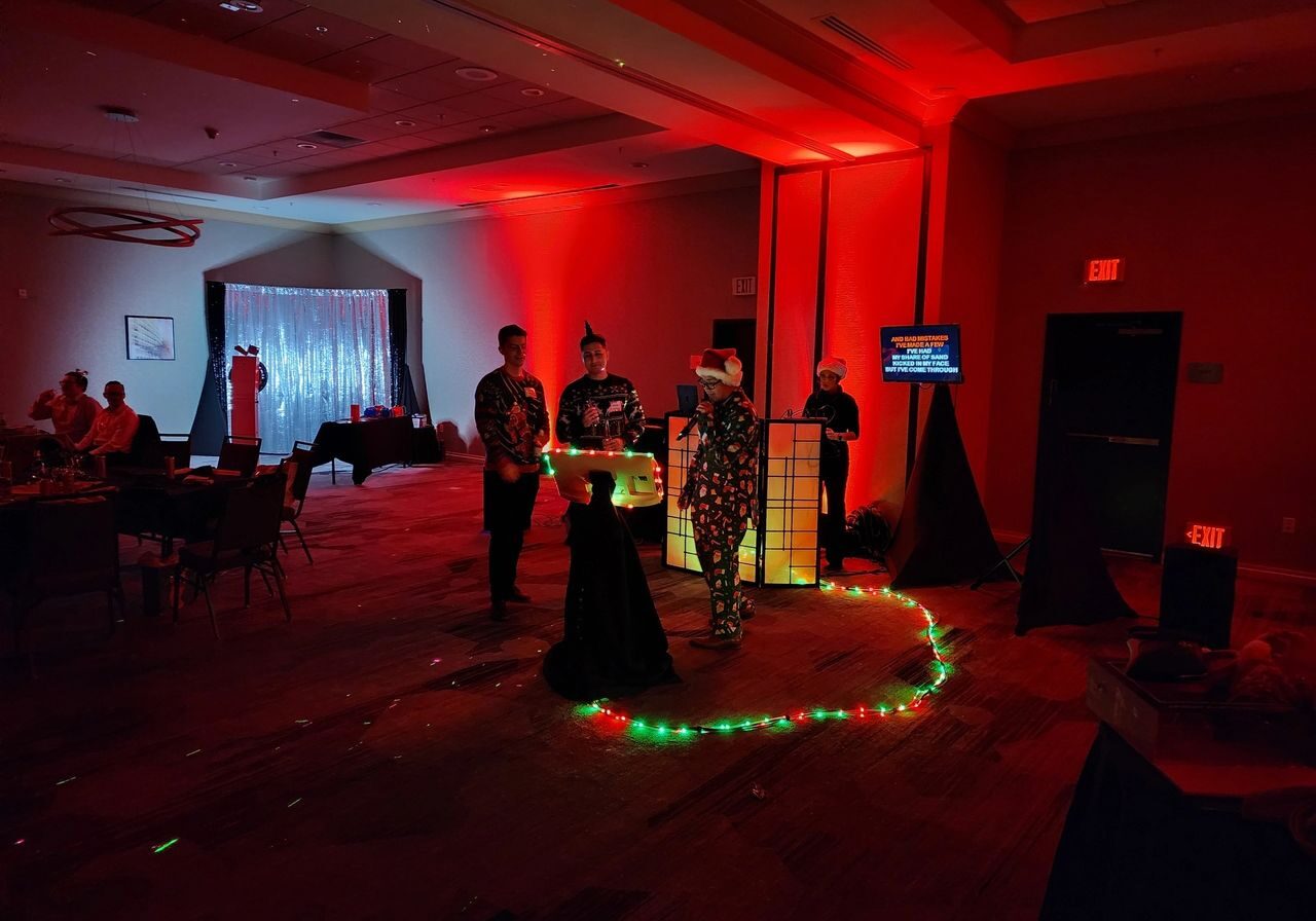 Holiday Party Christmas Four people in costumes stand near a lit DJ booth in a dimly lit room with red lighting and string lights on the floor; guests are seated at tables in the background.