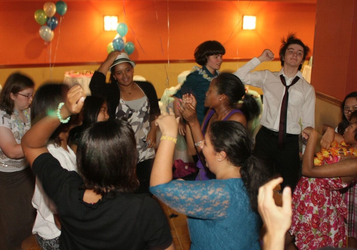 A group of people dance and celebrate together in a decorated room with balloons and orange walls.