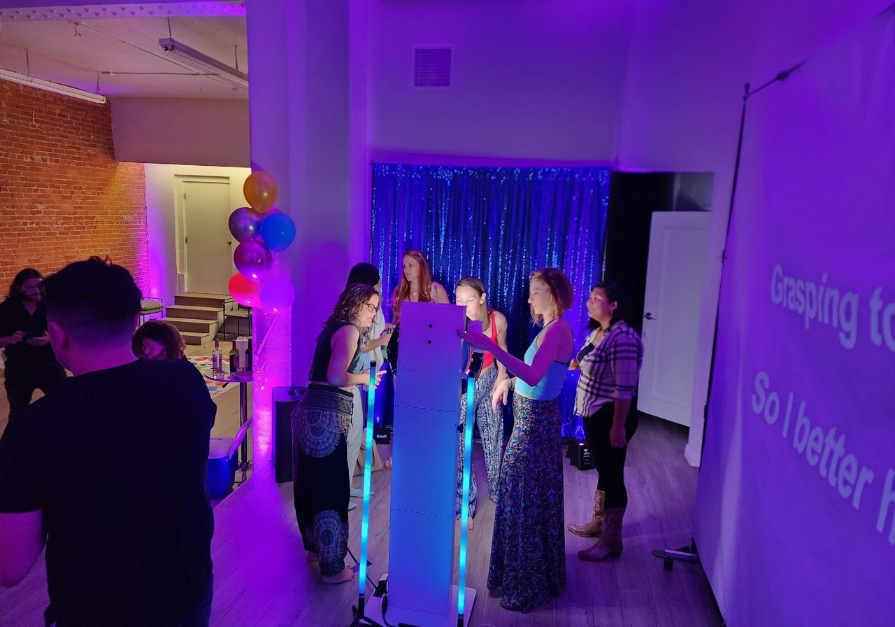 A group of people stand around a lighted photo booth at an indoor party with purple lighting; a karaoke screen is visible on the right.