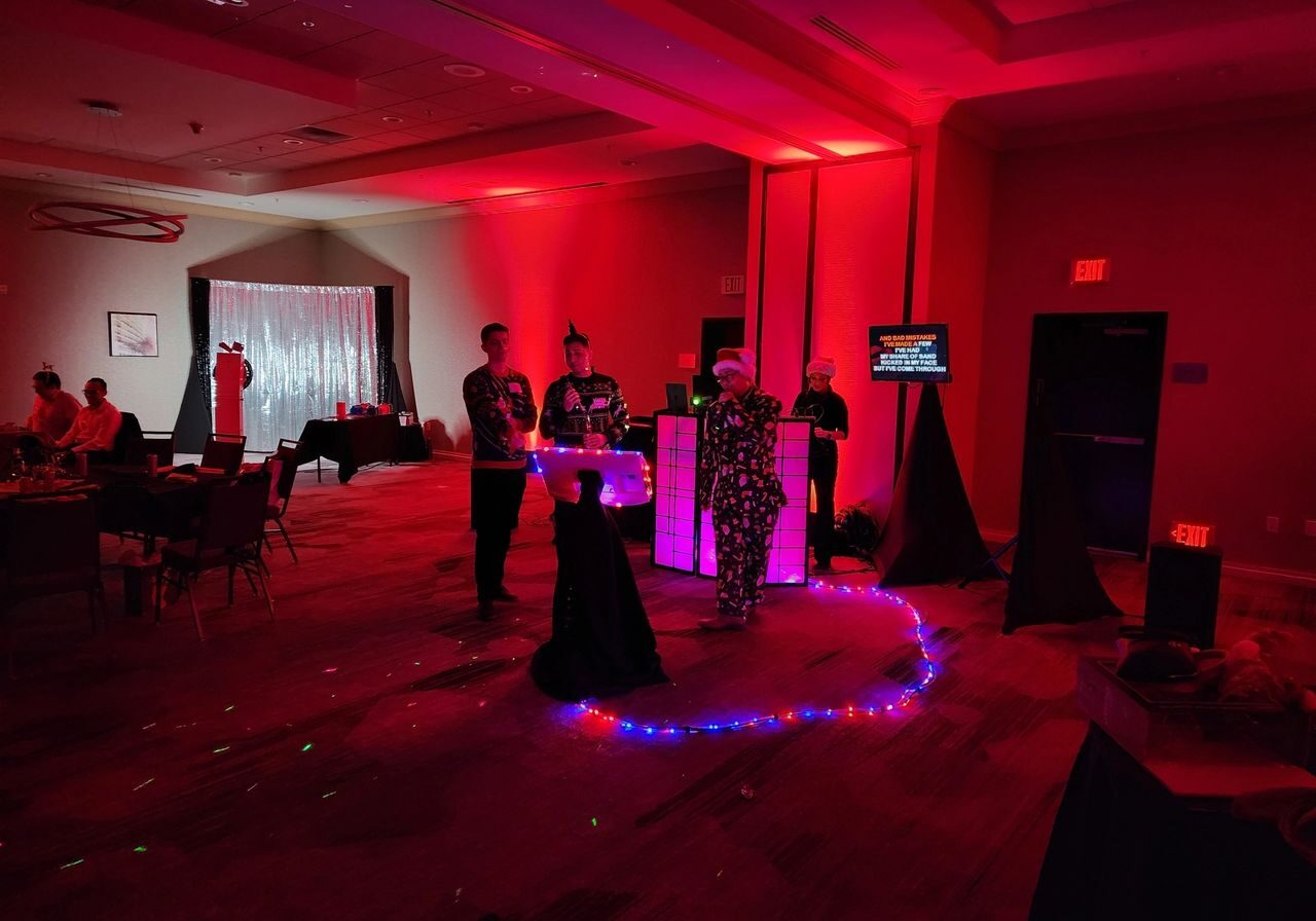 A group of people stands near a DJ booth with colorful lights in a dimly lit room decorated for a party or event.