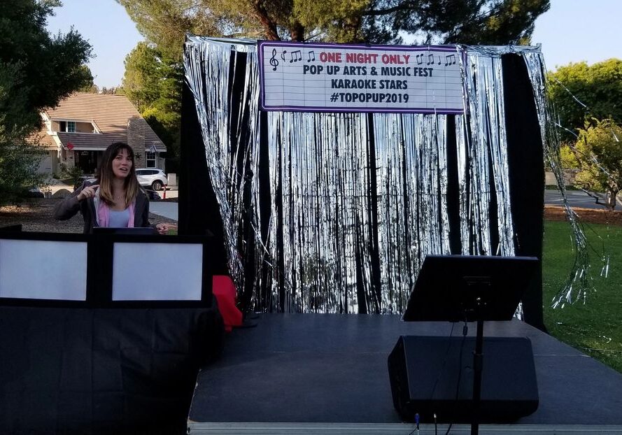 A woman stands behind a DJ booth on an outdoor stage decorated with silver streamers and a sign for a pop-up arts and music fest called “Karaoke Stars #TOPOPP2019.”.