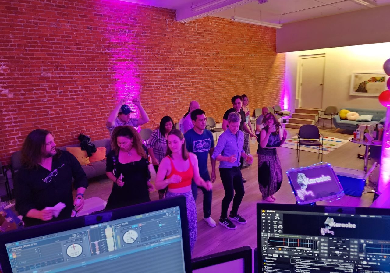 A group of people dance in a room with brick walls and purple lighting, viewed from behind a DJ booth with music equipment and karaoke screens.