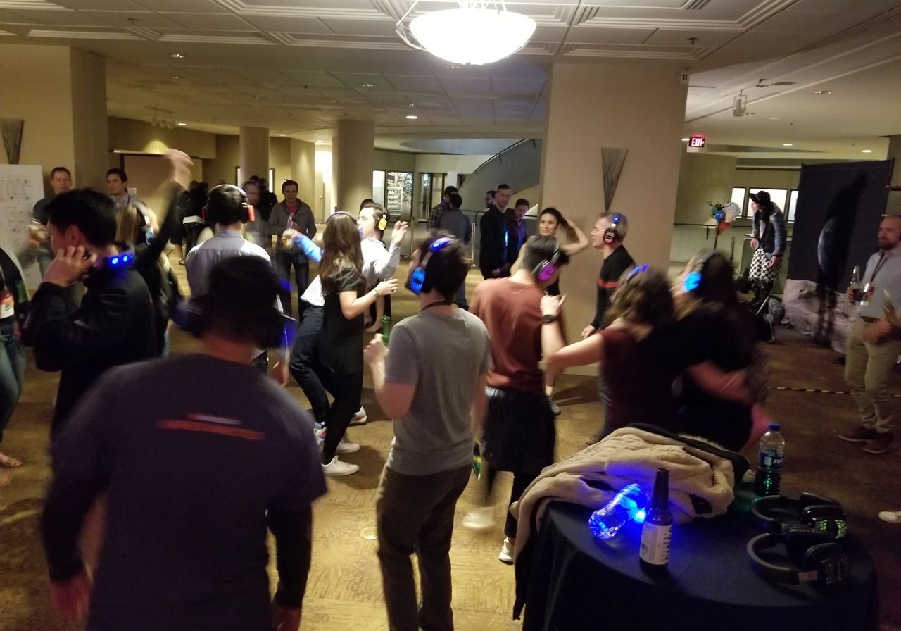 A group of people wearing headphones are dancing in a dimly lit room, likely participating in a silent disco event.