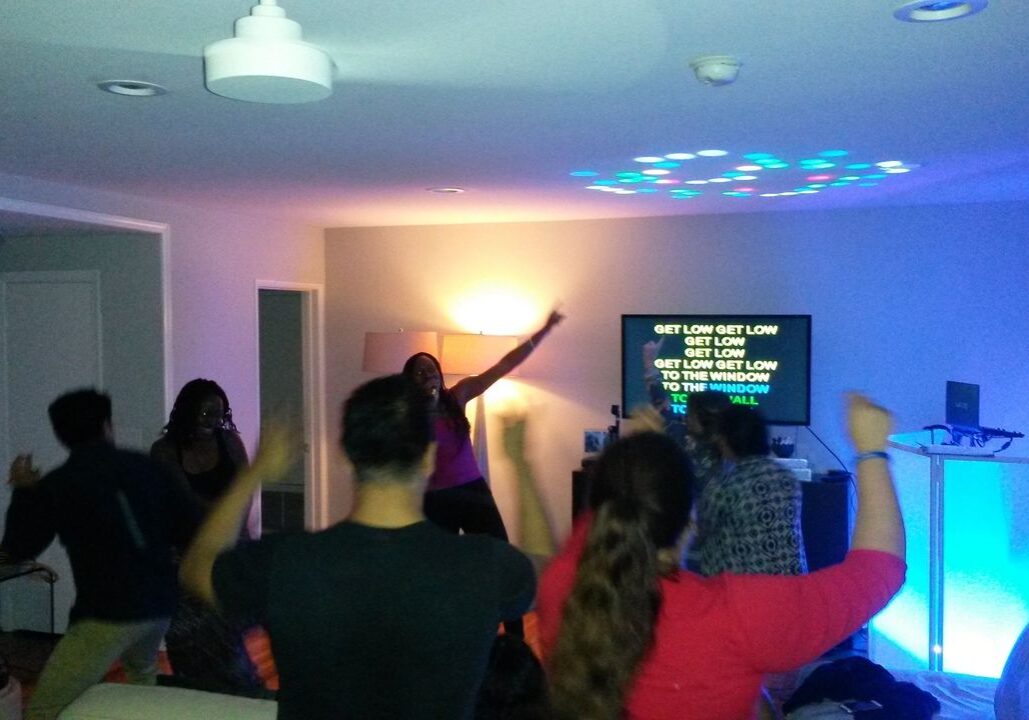 A group of people dance in a living room with dim lighting while karaoke lyrics are displayed on a TV screen in the background.