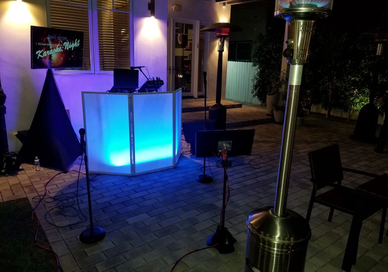 Outdoor karaoke setup with a glowing DJ booth, microphones, speakers, and patio heaters on a tiled patio at night. A "Karaoke Night" sign is visible in the background.