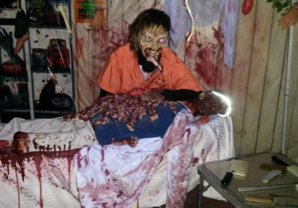 A zombie prop in an orange shirt appears to eat fake intestines from a bloodied mannequin on a table, surrounded by Halloween decorations and props.