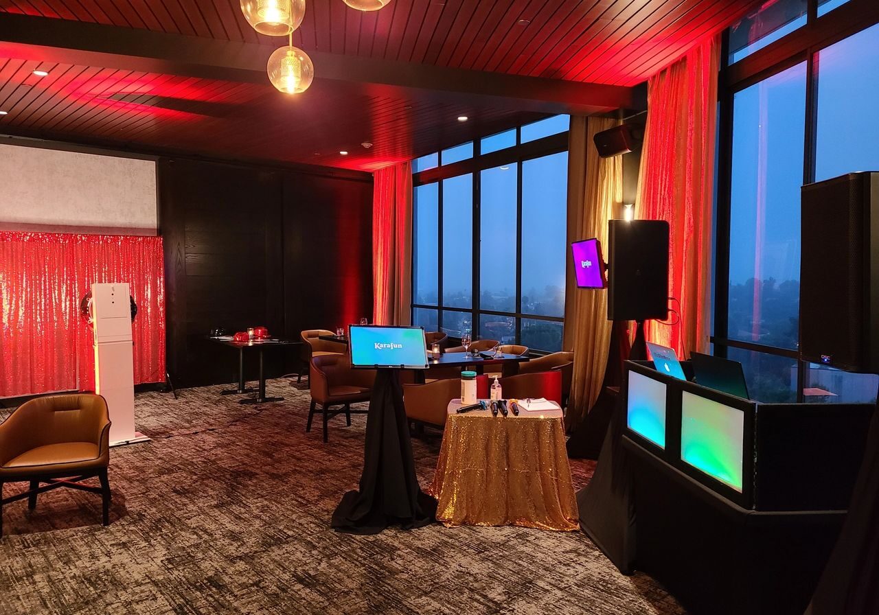 A modern event room with red lighting, lounge chairs, small tables, digital screens, a DJ setup, and large windows showing an evening cityscape.