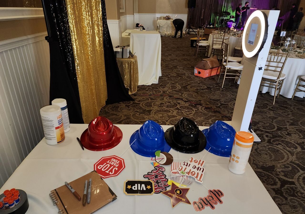 A table with colorful plastic hats, photo booth props, wipes, and a guest book, next to a ring light photo booth at an event venue with empty chairs and tables.