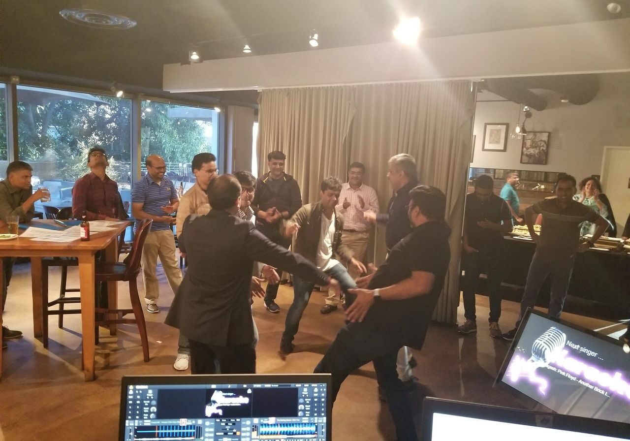 A group of people socialize and dance in a casual indoor setting, with tables, computers, and food visible in the background.