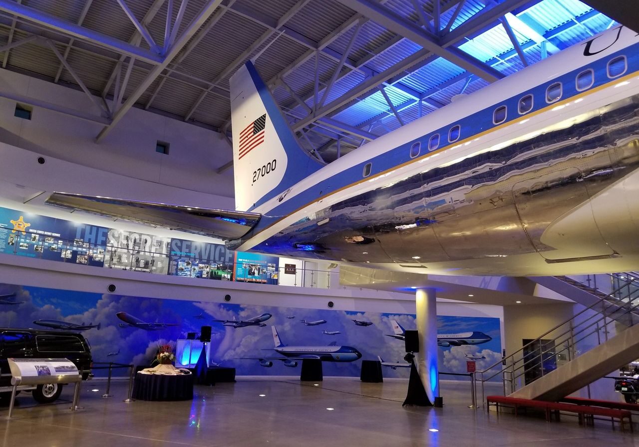 A polished aircraft with tail number 21000 and a U.S. flag is displayed indoors at a museum, with exhibits and murals of planes and Secret Service memorabilia in the background.