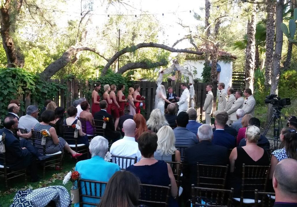 Outdoor wedding ceremony with a bride and groom at the altar, guests seated on chairs, and bridesmaids in red dresses and groomsmen in light suits. Trees and greenery in the background.