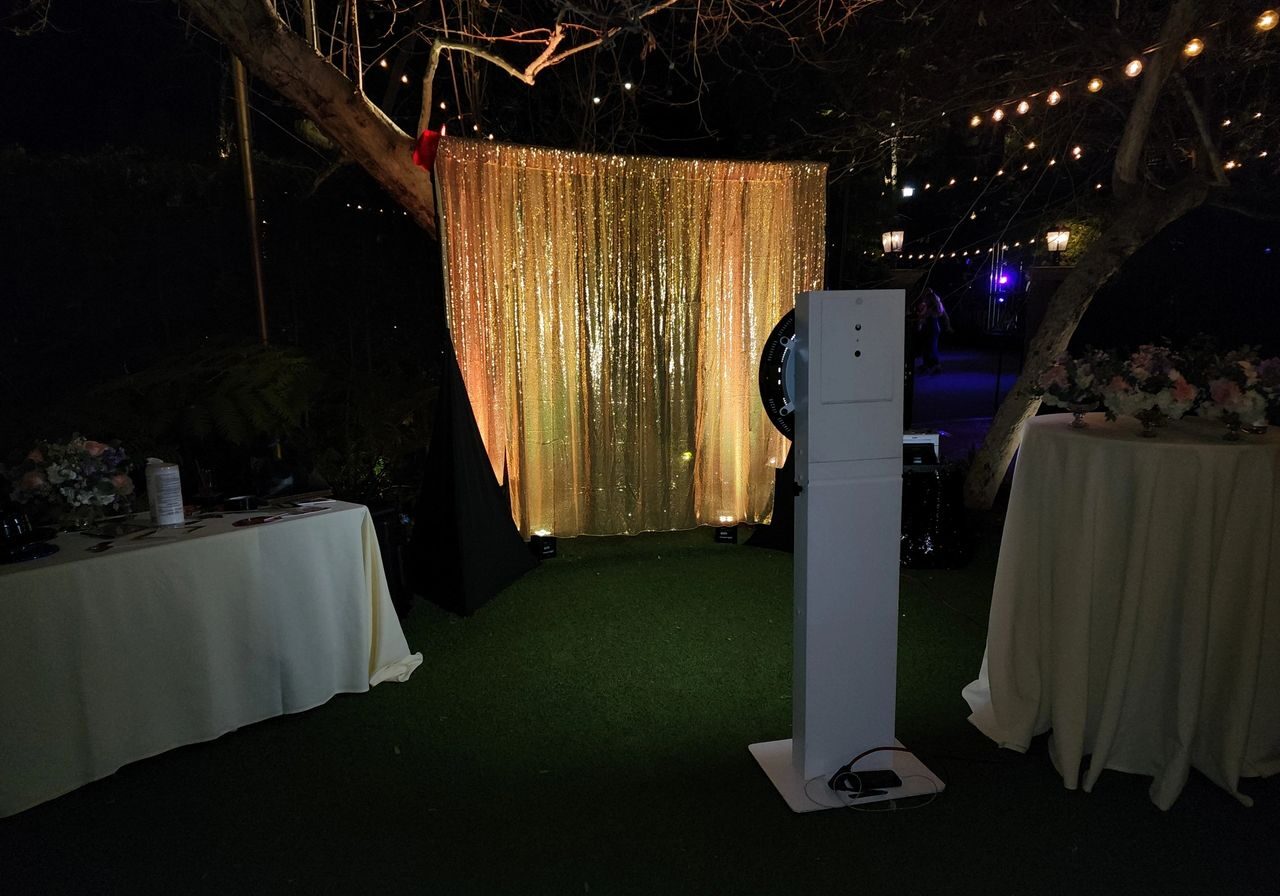 An outdoor photo booth setup with a gold sequin backdrop, a camera stand in front, and two round tables covered with white cloths and floral arrangements. String lights hang above.