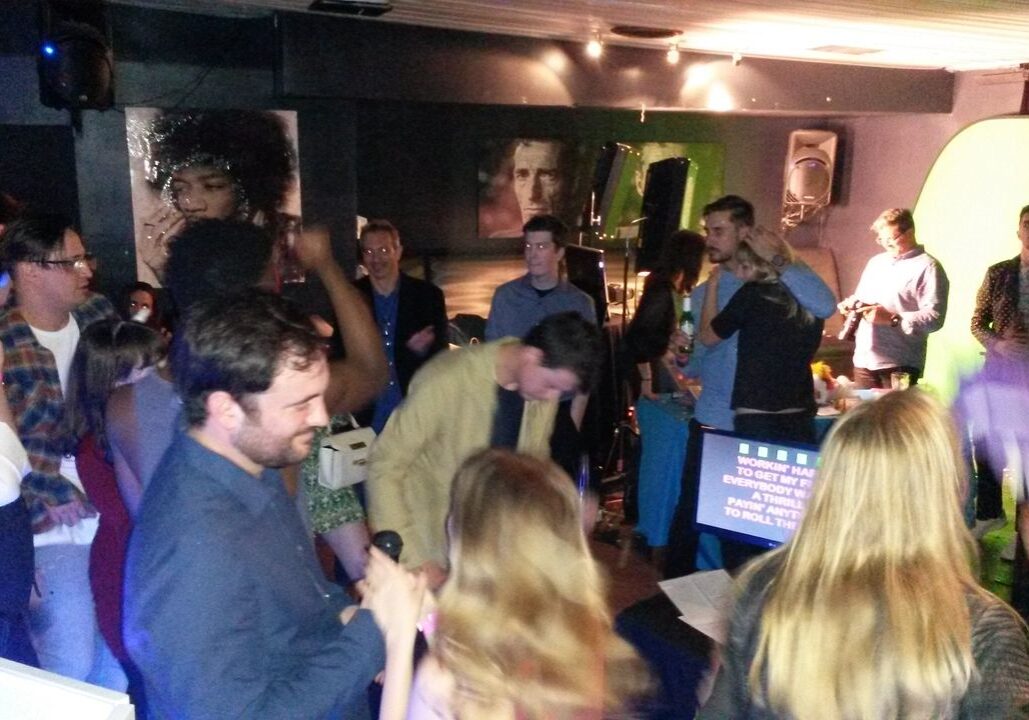 A group of people gather and socialize in a dimly lit room with a green screen and karaoke machine, while a few hold microphones and sing.