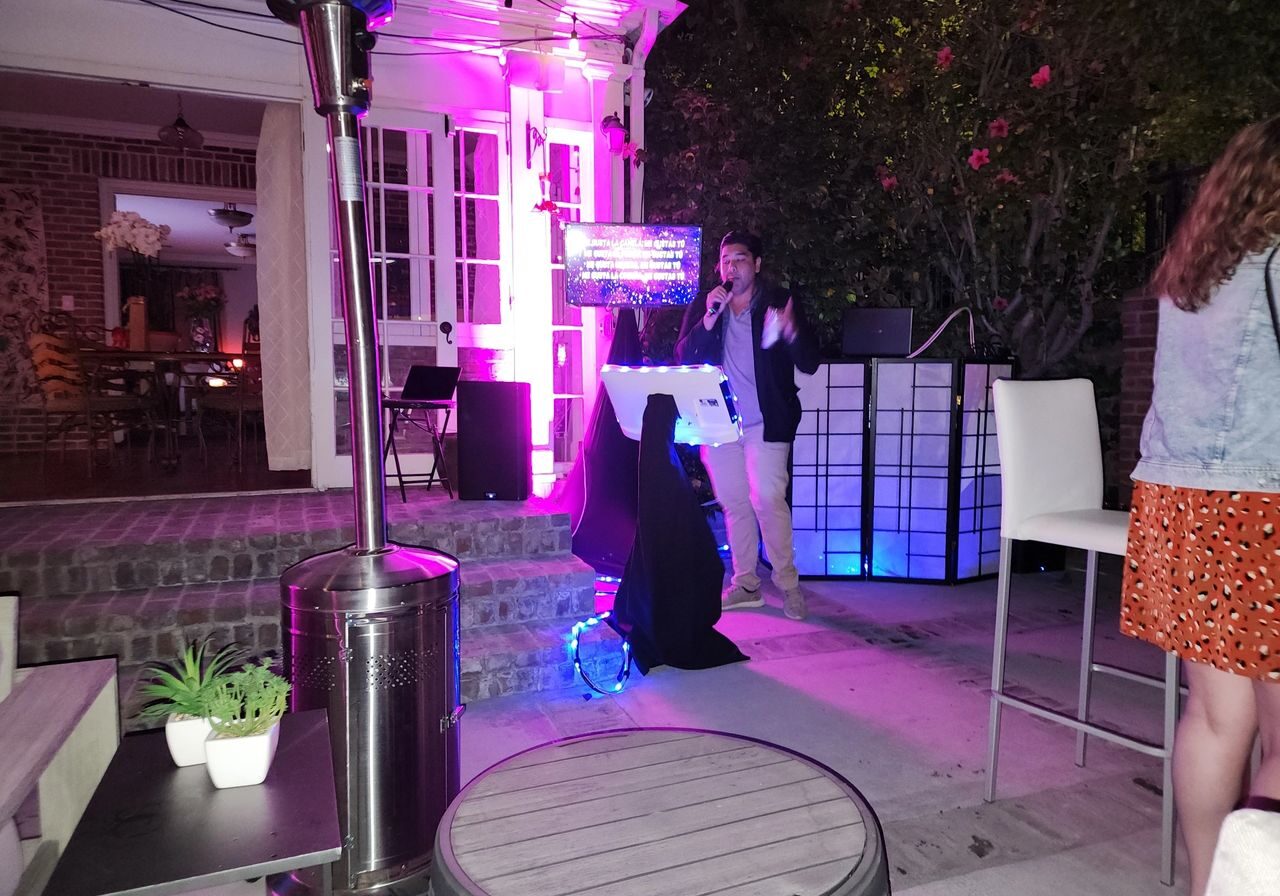 A person sings into a microphone at an outdoor nighttime event with colorful lights, audio equipment, and several people in the background.