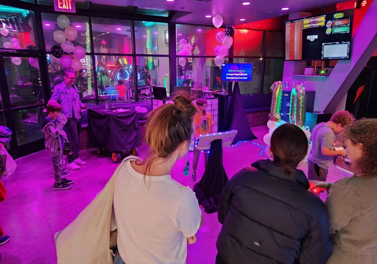 People gathered in a neon-lit room decorated with balloons; a child sings karaoke while others watch or interact with games and activities.