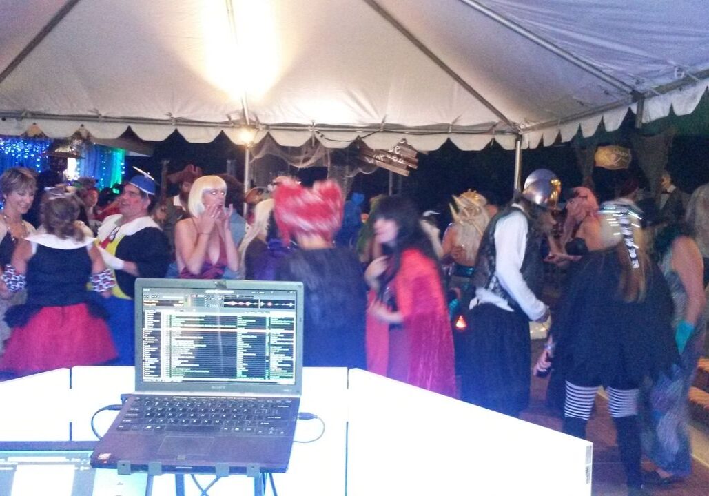 A group of people in costumes are dancing under a tent at a party, viewed from behind a DJ booth with a laptop and equipment.