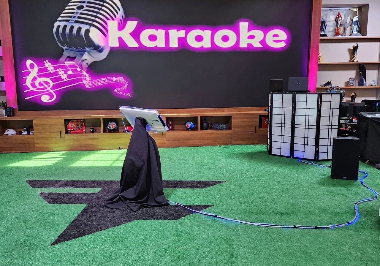 A karaoke setup with a microphone stand and screen on green turf in front of a large display that reads "Karaoke" with musical notes and a microphone graphic.