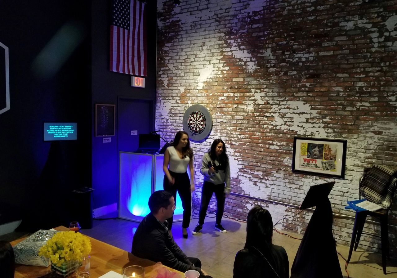 Two women perform karaoke in a dimly lit room with a brick wall, dartboard, and American flag; several people are seated and watching.