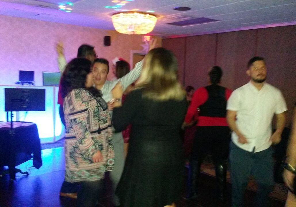 A group of people are dancing and socializing in a dimly lit room with a chandelier and a DJ setup in the background.