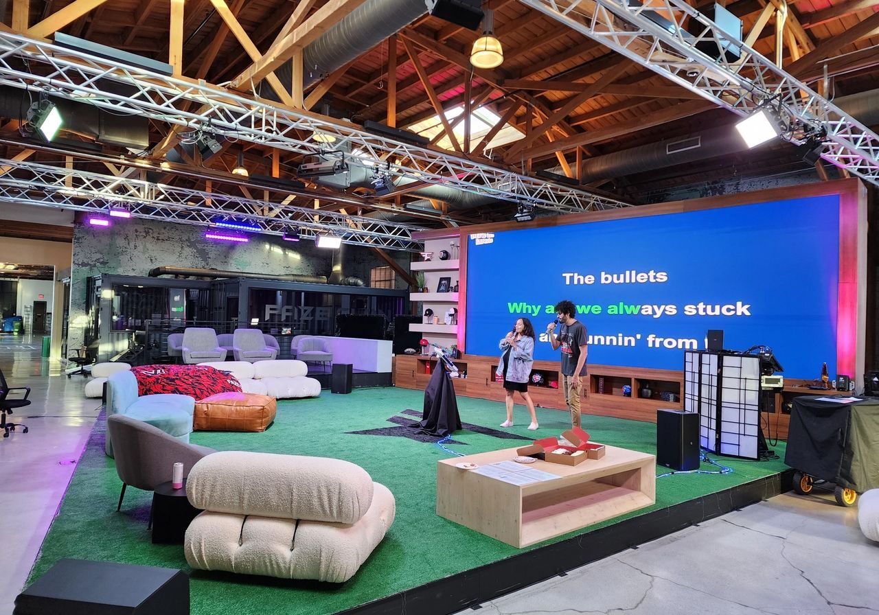 Two people stand on a stage with large karaoke lyrics displayed on a screen behind them in a spacious, modern, loft-style room with casual seating and exposed beams.