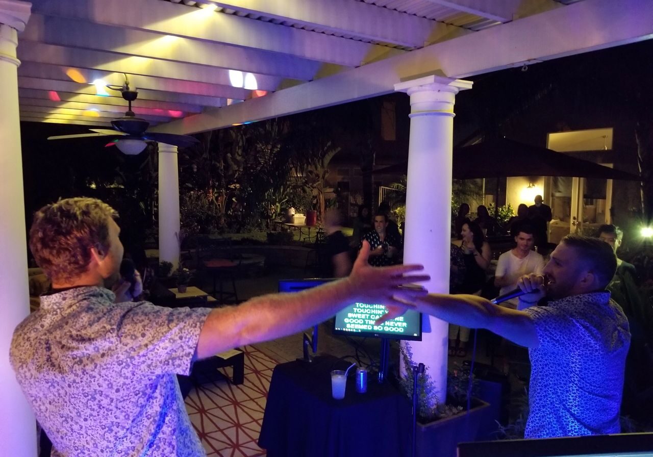 Two men sing karaoke with microphones on an outdoor patio at night, facing each other with arms outstretched, while people watch in the background.