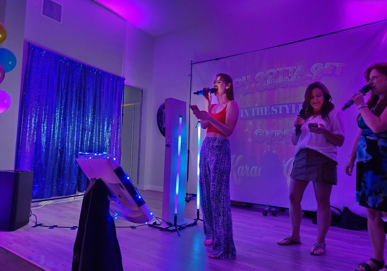Three women sing karaoke on a small stage with purple lighting, holding microphones. A screen behind them displays song lyrics. Balloons and a sequin curtain are visible to the left.
