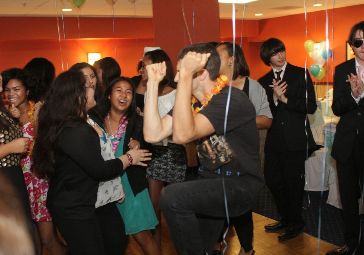 A group of young people at a party, some smiling and clapping, while a person in the center poses energetically with raised arms. Balloons and decorations fill the background, creating a lively scene with karaoke DJ fun.