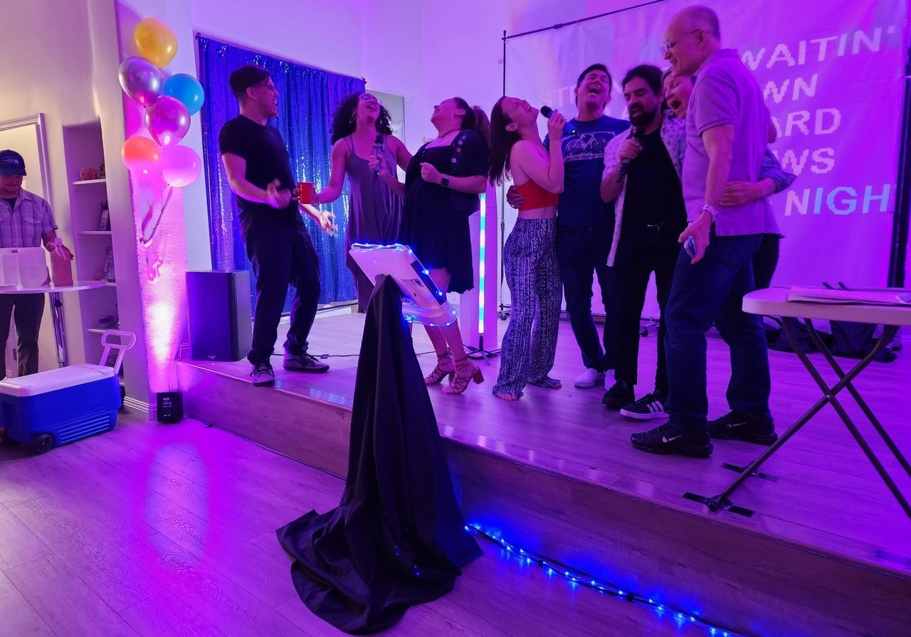 A group of people sing together on a small stage at a karaoke event, surrounded by colorful lights, balloons, and a projection screen in the background.