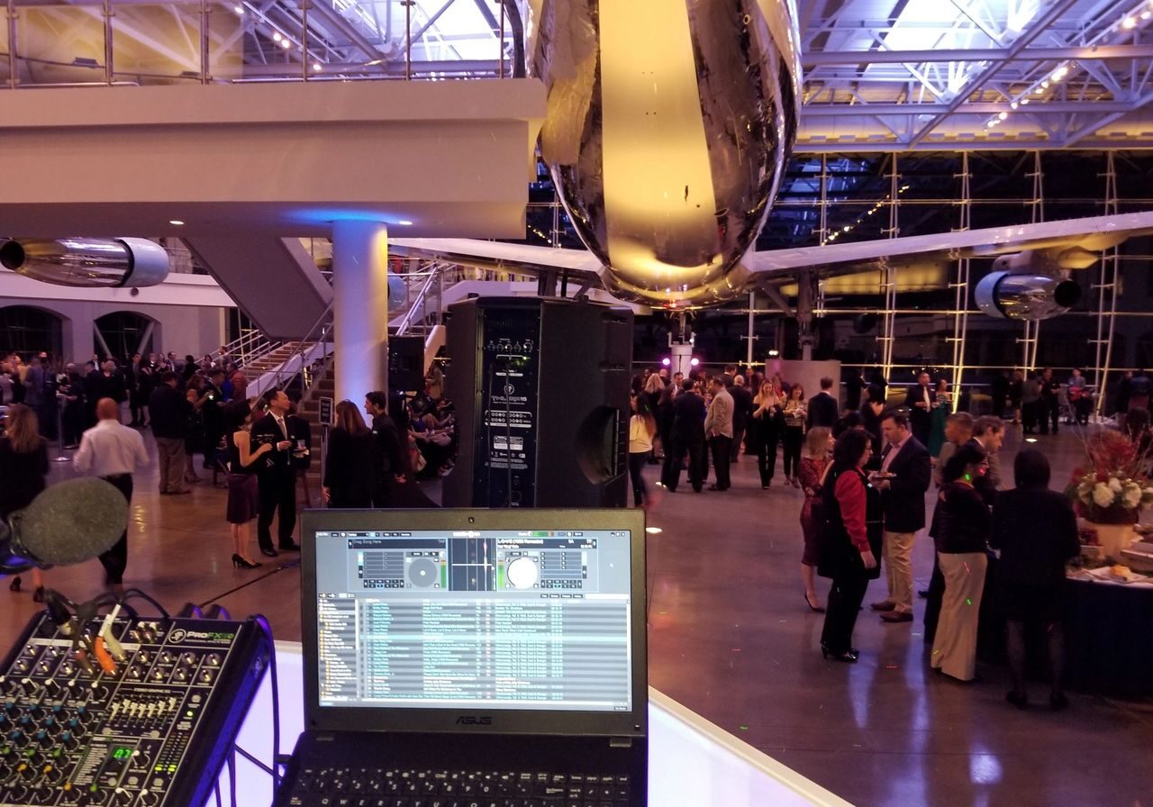 A DJ setup with a laptop and mixer overlooks a crowd of people mingling at an indoor event space with a large airplane suspended overhead.