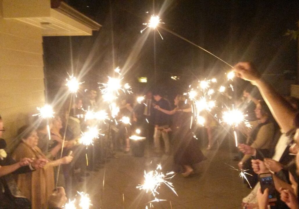 People form two lines holding sparklers at night while a couple dances in the middle of a pathway outside a building.