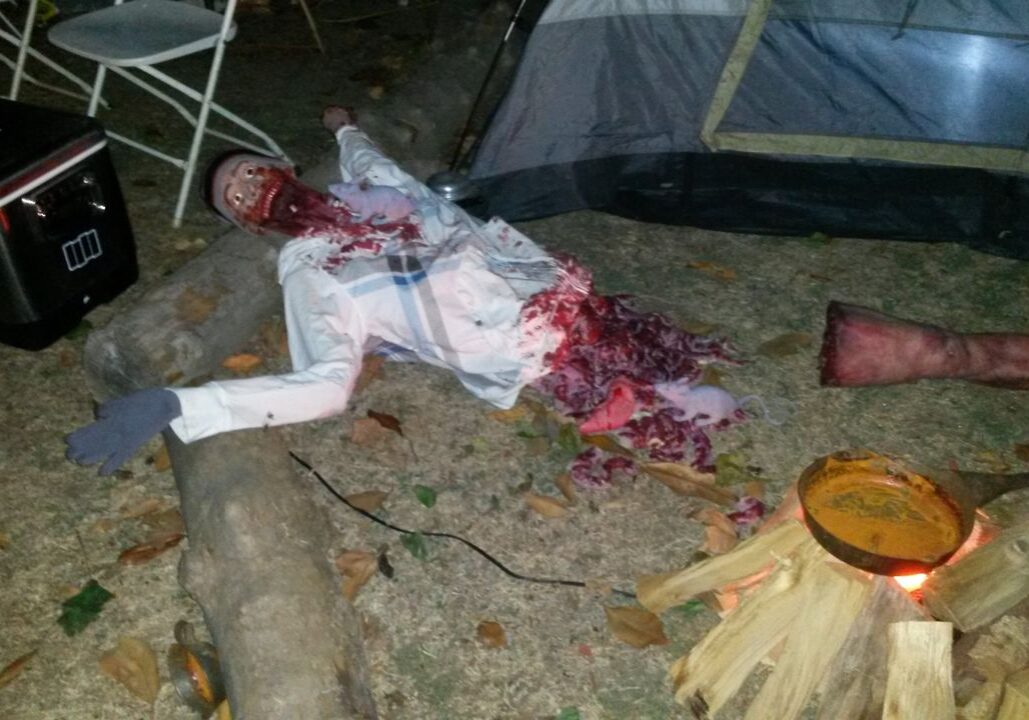 A Halloween prop of a mutilated body with fake blood and entrails is displayed on the ground near a tent and camping supplies.