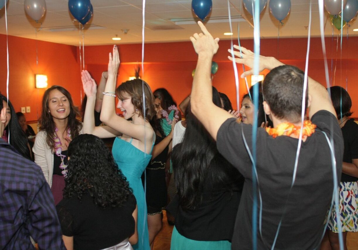A group of people are dancing and celebrating in a decorated indoor room with balloons and orange walls.