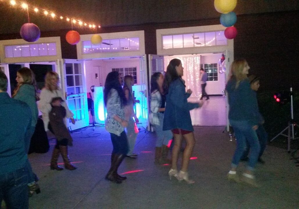 A group of people dance together on a patio at night under string lights and colorful paper lanterns, with a building and open doors in the background.