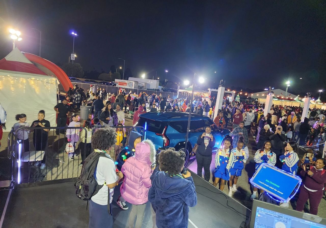 Large crowd at a nighttime outdoor event with tents, a blue car, performers on stage, and various people walking and gathering under bright lights.