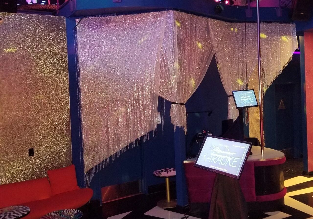 A brightly lit karaoke lounge with red seating, a stage, a karaoke machine, glittery decor, and a car displayed on a mezzanine level above.