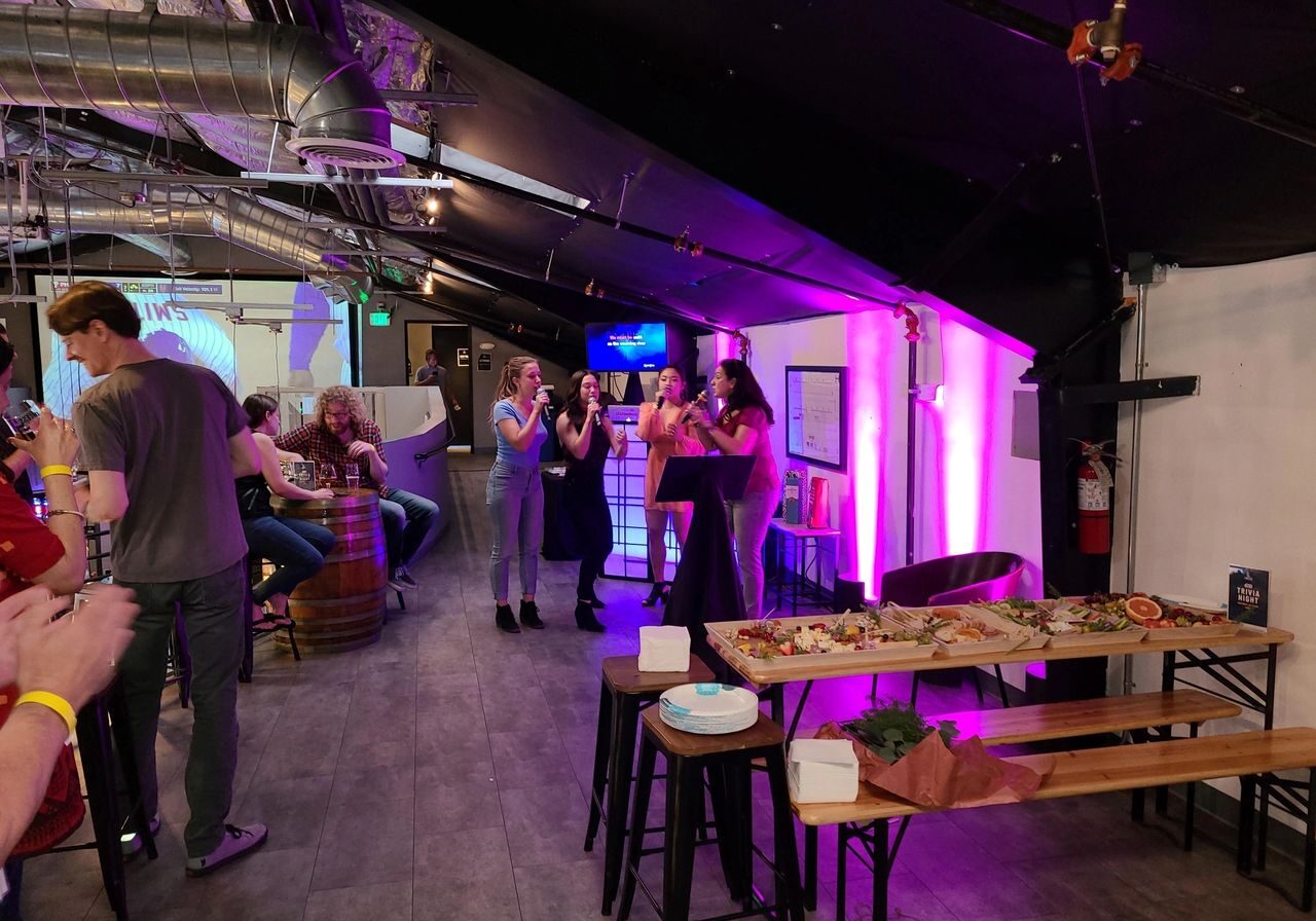 People socialize in a dimly lit venue with purple lighting; three women sing by a wall, and a table in the foreground holds food and plates.