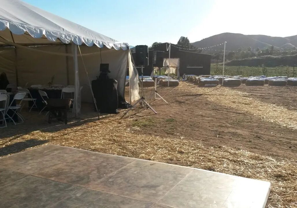 Outdoor event setup with a white tent containing chairs and tables on the left, a speaker and bales of hay on the right, and a wooden floor in the foreground, under a clear sky.