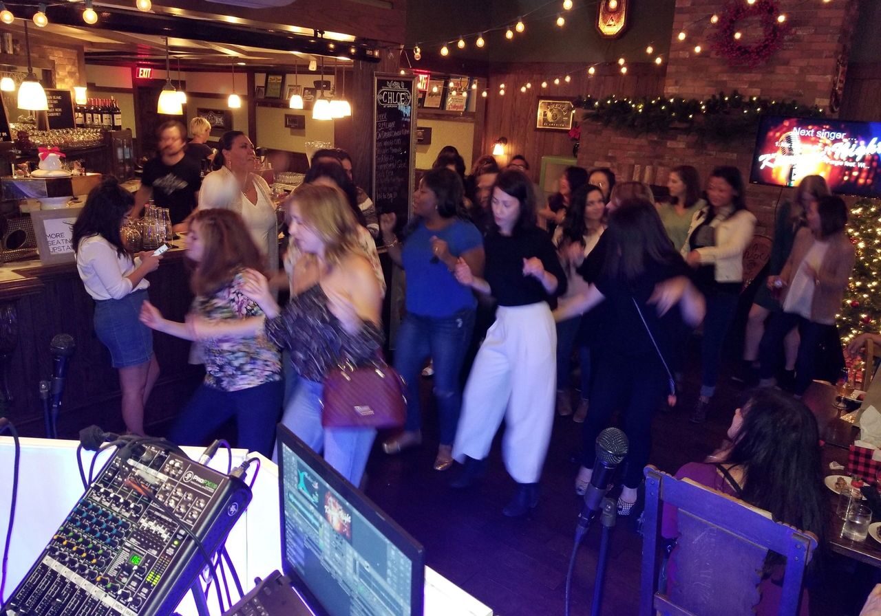 A group of people dance together in a warmly lit bar or restaurant, with music equipment visible in the foreground and festive decorations in the background.